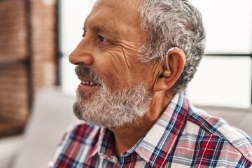 Senior grey-haired man using hearing aid sitting on sofa at home