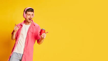 Fototapeta na wymiar Emotive young man in pink shirt posing in headphones with soda isolated over vivid yellow background. Flyer
