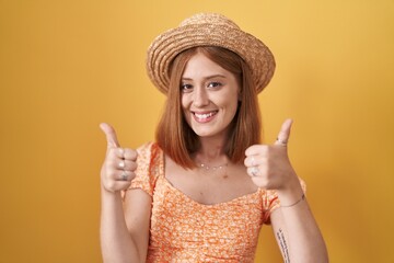 Young redhead woman standing over yellow background wearing summer hat success sign doing positive gesture with hand, thumbs up smiling and happy. cheerful expression and winner gesture.