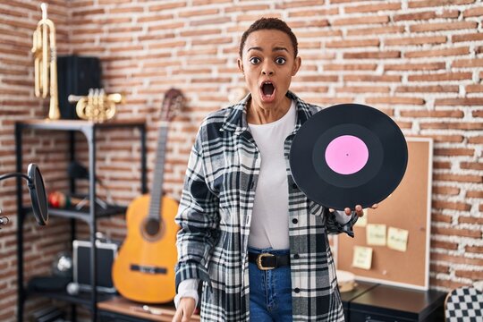 Beautiful african american woman holding vinyl record at music studio scared and amazed with open mouth for surprise, disbelief face