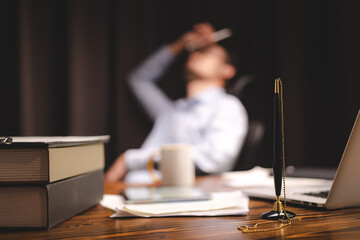 Closeup of a pen attached to a chain in stand lying on desk near laptop and books with documents with a business man sitting in blurry background in modern office cabin talking over mobile phone