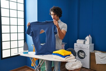 Hispanic man with curly hair ironing holding burned iron shirt at laundry room in shock face,...
