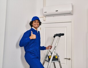 Hispanic man with curly hair working at home renovation smiling happy and positive, thumb up doing excellent and approval sign