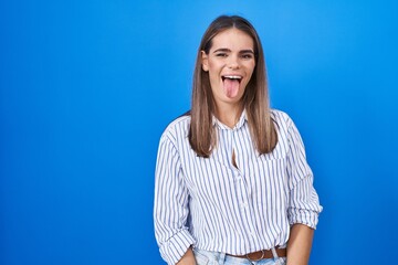 Hispanic young woman standing over blue background sticking tongue out happy with funny expression....