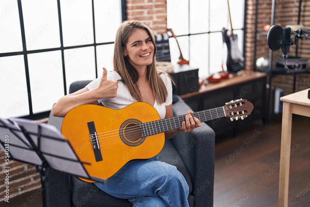 Poster Young beautiful woman playing classic guitar at music studio smiling happy and positive, thumb up doing excellent and approval sign