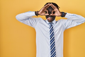 African man with dreadlocks standing over yellow background doing ok gesture like binoculars sticking tongue out, eyes looking through fingers. crazy expression.