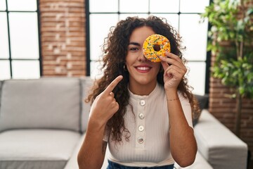 Young hispanic woman holding tasty colorful doughnut  eye smiling happy pointing with hand and...
