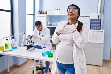 Young hispanic woman expecting a baby working at scientist laboratory smiling with hands on chest with closed eyes and grateful gesture on face. health concept.