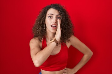 Hispanic woman with curly hair standing over red background hand on mouth telling secret rumor, whispering malicious talk conversation