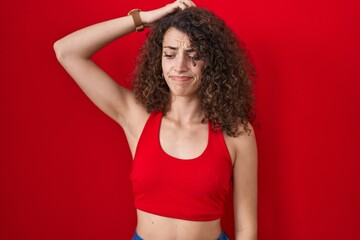 Fototapeta na wymiar Hispanic woman with curly hair standing over red background confuse and wondering about question. uncertain with doubt, thinking with hand on head. pensive concept.