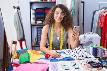 Fototapeta premium Young beautiful hispanic woman tailor using smartphone writing on notebook at clothing factory