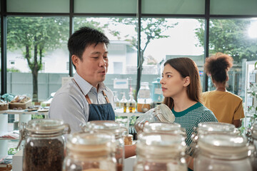 Asian male shopkeeper describes natural organic products to White woman customer in refill store, zero-waste and plastic-free grocery, environment-friendly, sustainable lifestyles in reusable shop.