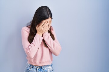 Young brunette woman standing over blue background with sad expression covering face with hands...