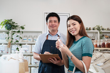 Asian male shopkeeper and female customer smile and look at camera at natural products in reusable containers at refill store, zero-waste groceries, organic retail, and sustainable shopping lifestyle.