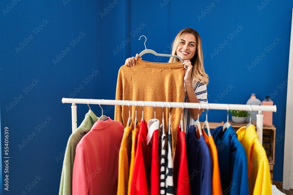 Wall mural young blonde woman smiling confident holding clothes of rack at laundry room - Wall murals