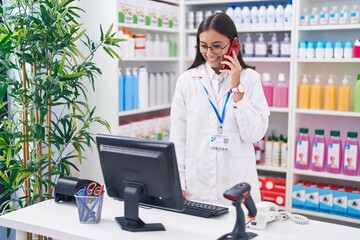 Young beautiful hispanic woman pharmacist talking on smartphone using computer at pharmacy