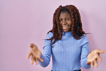 African woman standing over pink background smiling cheerful offering hands giving assistance and acceptance.