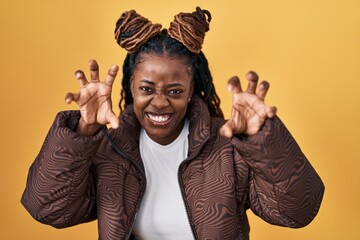 African woman with braided hair standing over yellow background smiling funny doing claw gesture as...