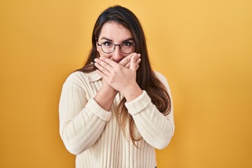 Young hispanic woman standing over yellow background shocked covering mouth with hands for mistake....