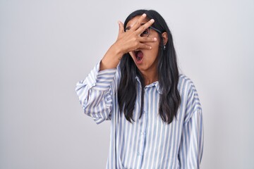 Young hispanic woman wearing glasses peeking in shock covering face and eyes with hand, looking through fingers with embarrassed expression.