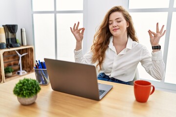 Young caucasian woman working at the office using computer laptop relaxed and smiling with eyes closed doing meditation gesture with fingers. yoga concept.