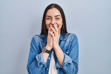Hispanic woman standing over blue background laughing and embarrassed giggle covering mouth with...