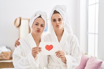 Middle age woman and daughter wearing bath robe holding heart card looking at the camera blowing a kiss being lovely and sexy. love expression. - obrazy, fototapety, plakaty