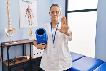 Young physiotherapist woman holding foam roll at the clinic with open hand doing stop sign with serious and confident expression, defense gesture