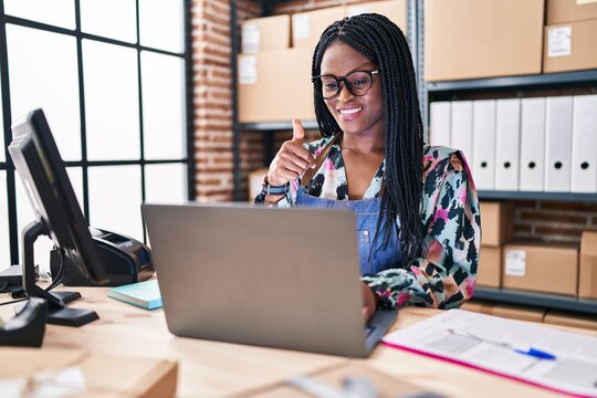 African woman with braids working at small business ecommerce with laptop smiling happy and positive, thumb up doing excellent and approval sign