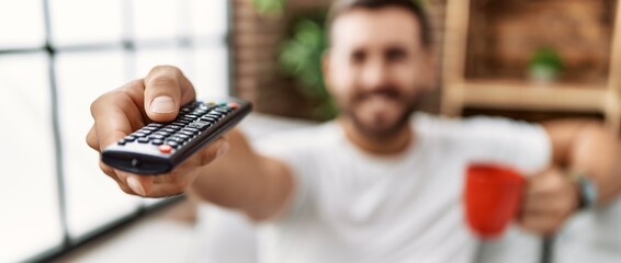 Young hispanic man drinking coffee watching tv at home