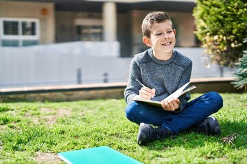 Blond child writing on book sitting on grass at park