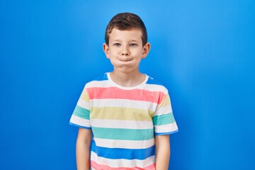 Young caucasian kid standing over blue background puffing cheeks with funny face. mouth inflated with air, crazy expression.