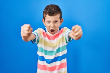 Young caucasian kid standing over blue background angry and mad raising fists frustrated and furious while shouting with anger. rage and aggressive concept.