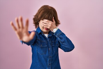 Middle age woman standing over pink background covering eyes with hands and doing stop gesture with...