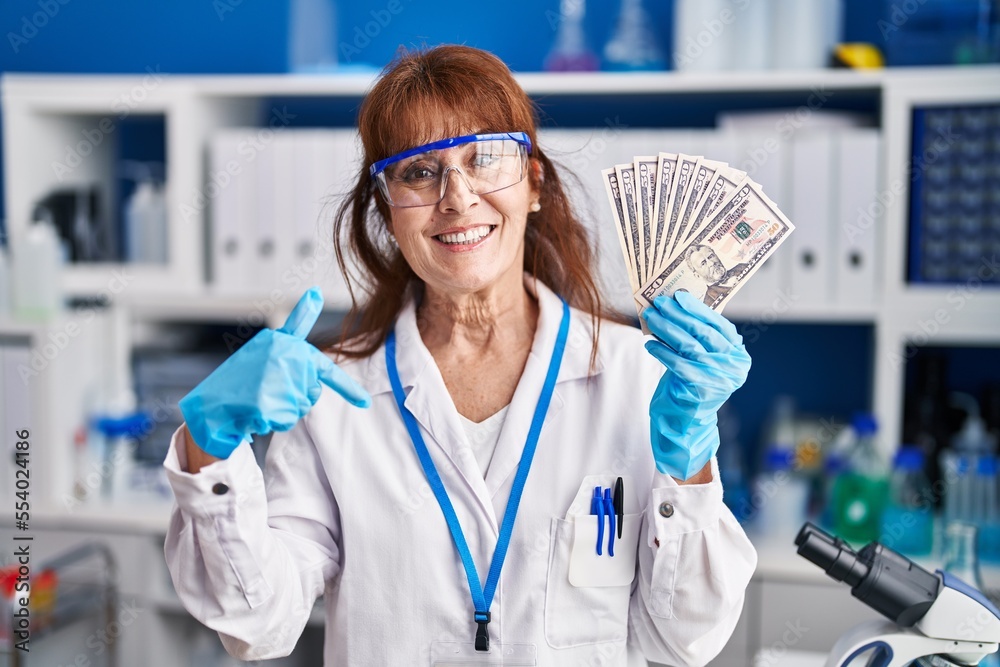 Wall mural middle age hispanic woman working at scientist laboratory holding dollars pointing finger to one sel