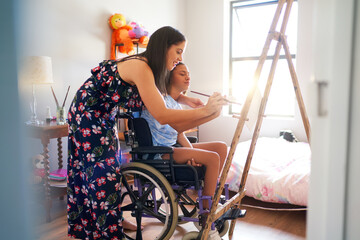Mother and disabled daughter in wheelchair painting in bedroom