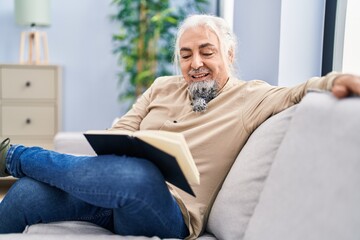 Middle age grey-haired man reading book sitting on sofa at home