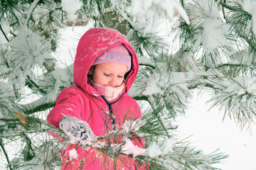 winter portrait of a cute beautiful girl, blonde 7-9 years old, in a bright overalls, standing near a pine tree covered with snow