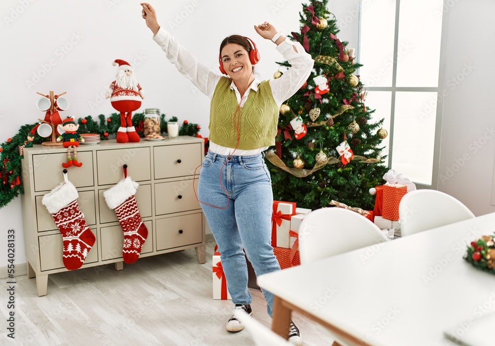 Sticker Young beautiful hispanic woman listening to music and dancing standing by christmas tree at home