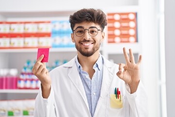 Arab man with beard working at pharmacy drugstore holding condom smiling looking to the side and...