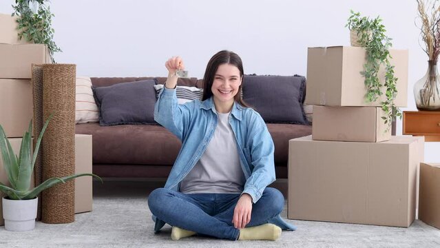 Happy young woman student showing the keys sitting on the floor in a to her new house. Smiling female rejoices in moving