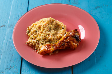 Couscous with vegetables in sauce and spices with grilled chicken drumstick on a plate on a blue table.