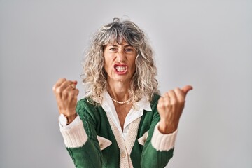 Middle age woman standing over white background angry and mad raising fists frustrated and furious...