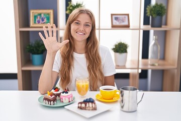 Young caucasian woman eating pastries t for breakfast showing and pointing up with fingers number five while smiling confident and happy.