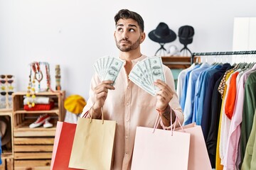 Young hispanic man holding shopping bags and dollars at retail shop smiling looking to the side and staring away thinking.