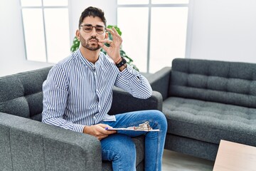 Young psychologist man at consultation office mouth and lips shut as zip with fingers. secret and...
