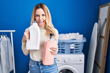 Young caucasian doctor woman holding detergent bottle at laundry room sticking tongue out happy with funny expression.