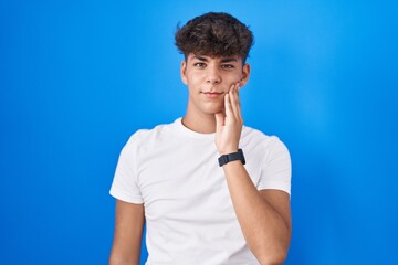 Hispanic teenager standing over blue background touching mouth with hand with painful expression because of toothache or dental illness on teeth. dentist