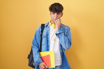 Hispanic teenager wearing student backpack and holding books looking stressed and nervous with hands on mouth biting nails. anxiety problem.