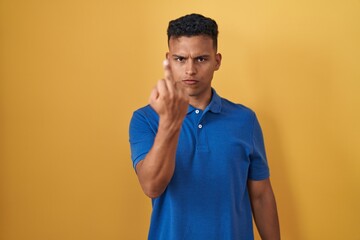 Young hispanic man standing over yellow background showing middle finger, impolite and rude fuck...